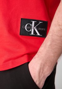 Czerwona bawełniana koszulka z krótkim rękawem, z czarną prostokątną naszywką z białym logiem "CK" oraz tekstem "Calvin Klein Jeans". Prosty dół.