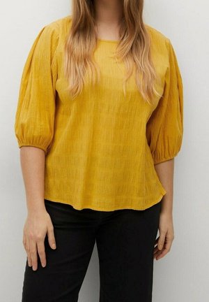 Femme aux longs cheveux blonds portant une blouse jaune moutarde texturée avec des manches bouffantes mi-coudes et un pantalon noir, sur un fond uni.