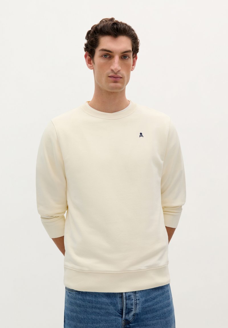 Cremefarvet sweatshirt lavet af blødt stof, med rund hals, ribbede manchetter og et lille mørkeblåt kranielogo på brystet.