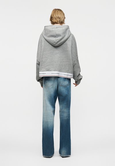 Άτομο με κοντά ξανθά μαλλιά που φοράει φαρδιά γκρι μπλούζα με κουκούλα και διακοσμητική ταινία "DSQUARED2" και ξεθωριασμένο φαρδύ τζιν, όρθιο με την πλάτη προς τα εμάς.