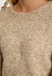 Bežani pleteni pulover s teksturiranim, pahuljastim završetkom i okruglim izrezom. Tkanina izgleda mekano s krupnim uzorkom tkanja.