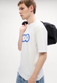Hvid bomulds T-shirt med et blåt og hvidt grafisk tekstdesign. Afslappet pasform, korte ærmer, suppleret med en sort rygsæk.