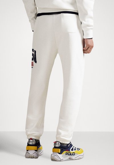 Biele tepláky s námorníckymi akcentmi, s zadným vreckom a výrazným nápisom "USA". Osoba má na nohách viacfarebné tenisky s logom značky.