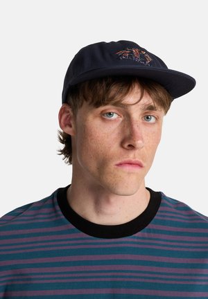Gorra azul marino con bordado frontal, visera negra, usada con una camiseta de manga corta a rayas en tonos verde azulado y púrpura, con un cuello negro.