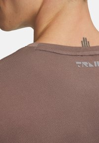 Camisa corta de manga corta texturizada en marrón con cuello redondo y un sutil detalle del logo en la parte posterior. Tejido suave con un diseño transpirable.