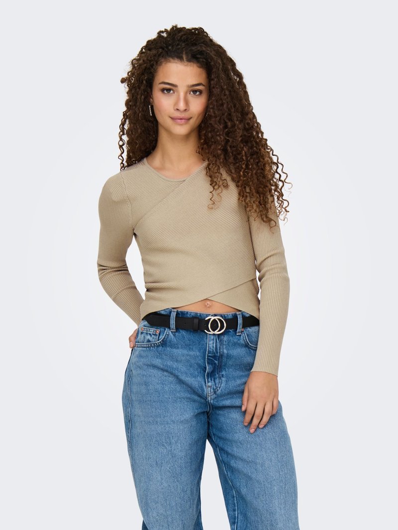Giovane donna con capelli lunghi e ricci che indossa un top beige a coste con maniche lunghe e jeans blu a vita alta con cintura nera, in piedi con la mano in tasca.