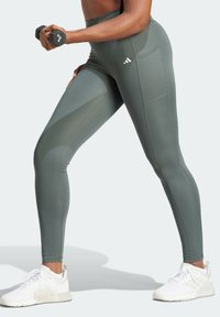 Leggings a vita alta di colore verde oliva realizzati in tessuto elastico, dotati di tasche laterali e cuciture a contrasto, abbinati a scarpe da ginnastica bianche.