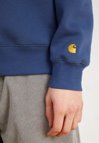 Zils sporta krekls ar ribinātām piedurknēm, uz kurām ir neliels dzeltenais logotips. Audums izskatās mīksts un ar faktūru, apvienots ar pelēkiem kordurojā izgatavotiem biksēm.