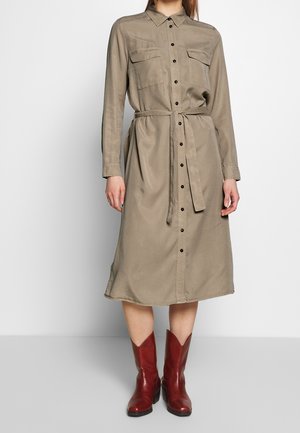 Mujer con vestido camisa beige de manga larga hasta la rodilla, con botones y cinturón anudado, combinado con botas de cuero rojas hasta media pantorrilla.