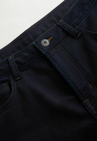 Mørkeblå denimjeans med knaplukning, bæltestropper og synlige syninger. Stoffet har en glat tekstur med let glans.