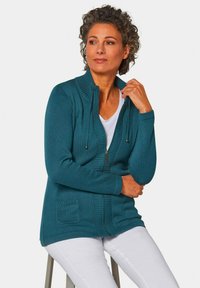 Türazu Zip-up Cardigan aus strukturiertem Strickstoff, mit hohem Kragen, Kordelzügen und einer frontalen Tasche. Getragen über einem weißen Oberteil und Hose.