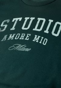 Tissu vert foncé avec texte blanc indiquant "STUDIO," "AMORE MIO," et "Milano" en différentes polices et tailles.