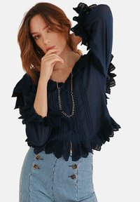 Navyblaues Bluse mit Spitze, Rüschensäumen und Knopfdetails. Kombiniert mit gestreiften, hochtaillierten Hosen mit braunen Knöpfen.