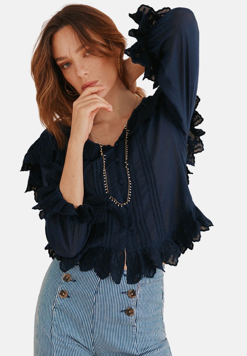Navyblaues Bluse mit Spitze, Rüschensäumen und Knopfdetails. Kombiniert mit gestreiften, hochtaillierten Hosen mit braunen Knöpfen.