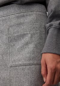 Un tissu en feutre gris capture les détails de texture, avec une grande poche brodée et une manche à poignets en tricot gris assorti.