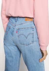 Jeans de ganga azul claro com cintura alta, apresentando costura laranja contrastante, um emblema de couro e design de bolso traseiro característico.