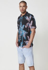 Camisa negra de manga corta con un patrón de palmeras en rosa y azul, combinada con pantalones cortos de mezclilla claros. Tejido suave y corte relajado.
