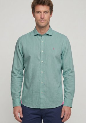 Hombre que lleva una camisa de manga larga color azul verdoso con un logo bordado y pantalones azul marino, de pie sobre un fondo claro y liso.