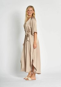 Langes beiges Kleid mit einem Taillengürtel, kurzen Ärmeln und seitlichen Schlitzen. Hergestellt aus einem glatten, fließenden Stoff. Kombiniert mit hellen Sandalen.