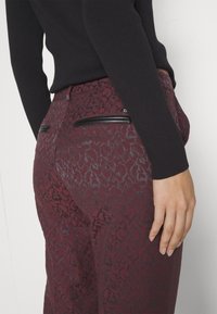 Οίνοπα παντελόνι με υφή ζωικής εκτύπωσης, διαθέτοντας μαύρες τσέπες με φερμουάρ και εφαρμοστή σχεδίαση, συνδυασμένο με μαύρη μπλούζα.