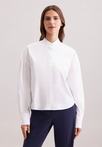 Camicia bianca a maniche lunghe con colletto rialzato e patta dei bottoni. Tessuto morbido con una superficie liscia. Indossata con pantaloni blu navy.
