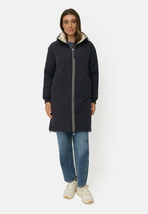 Femme portant une longue doudoune noire avec une capuche beige, un jean bleu et des baskets blanches, debout devant un fond uni.