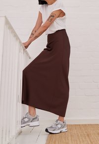 Gonna maxi marrone realizzato in un materiale liscio, abbinato a scarpe da ginnastica grigie. La persona è appoggiata a una ringhiera bianca.
