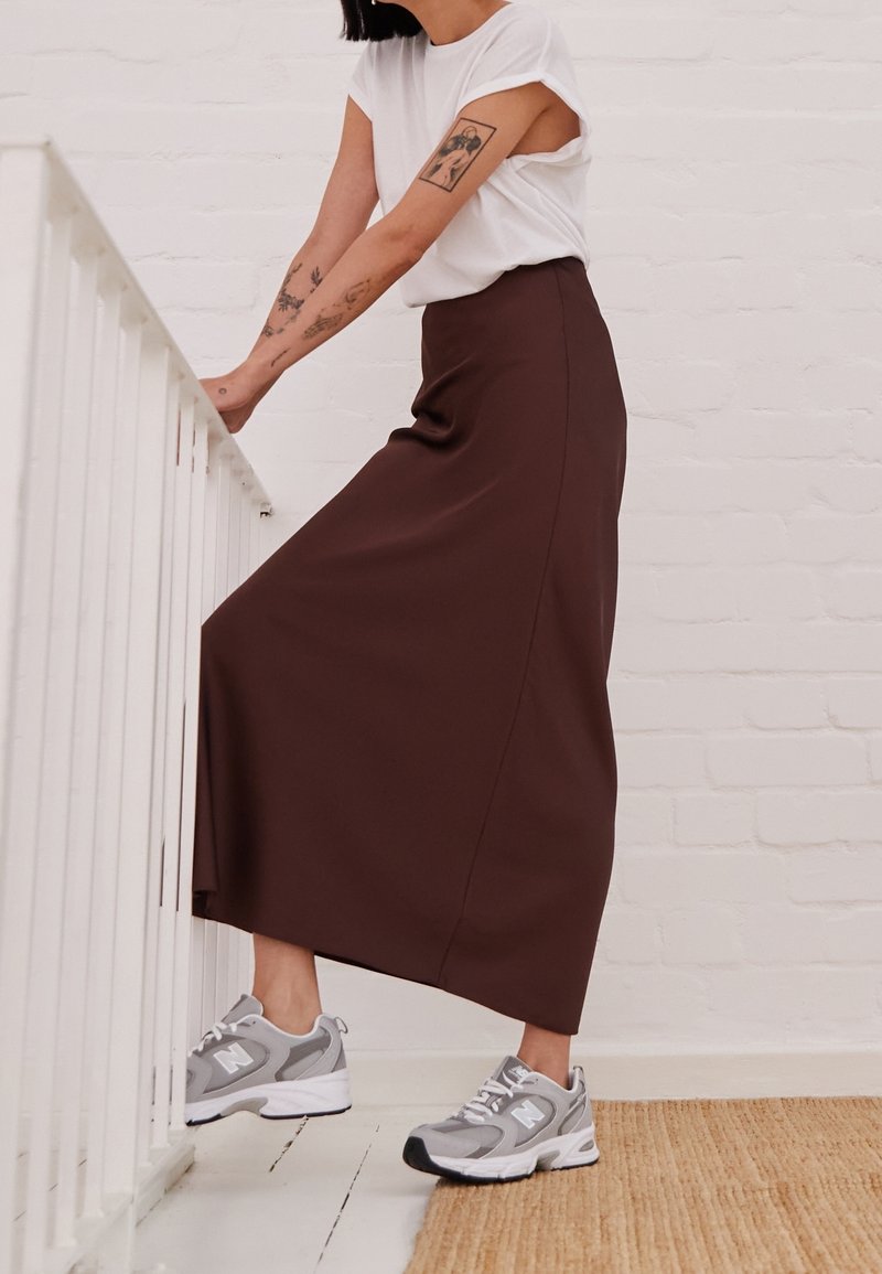 Gonna maxi marrone realizzato in un materiale liscio, abbinato a scarpe da ginnastica grigie. La persona è appoggiata a una ringhiera bianca.