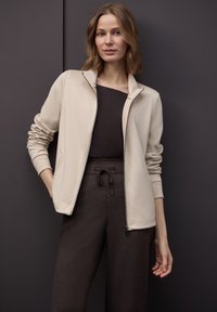 Giacca beige con zip e collo alto, indossata sopra una maglia marrone, abbinata a pantaloni marrone scuro con texture e vita con coulisse.