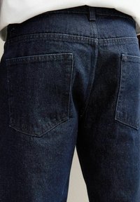 Los jeans de mezclilla oscura presentan un diseño clásico con dos bolsillos traseros, un corte recto y un acabado ligeramente texturizado. El color es un azul marino profundo.