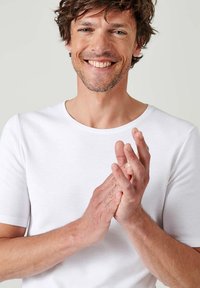 T-shirt bianca di cotone con scollo a girocollo, maniche corte e design aderente. Il modello sta applaudendo, mettendo in mostra la pelle liscia e i capelli mossi.