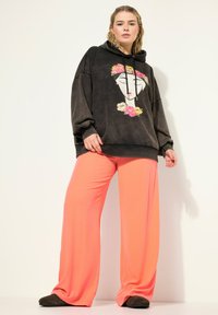Černá mikina s kapucí a květinovým grafikou na obličeji, kombinovaná s jasně korálovými kalhotami s širokým střihem. Měkká látka, uvolněný střih a hladký povrch.