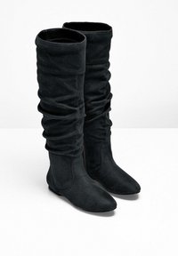 Bottes noires en daim montantes avec un design décontracté, semelles plates et bouts pointus, se tenant côte à côte sur une surface blanche.