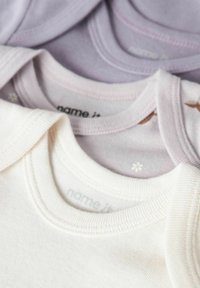 Trois hauts pour bébé empilés : un crème, un lavande clair avec un imprimé floral subtil, et un lavande pâle. Tous présentent des encolures côtelées et un tissu doux.