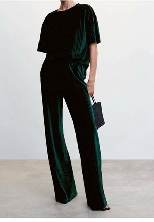 Personne portant un pantalon large plissé vert foncé et un haut à manches courtes assorti, tenant un petit sac à main noir, debout sur un sol neutre.