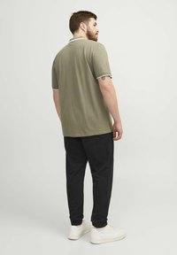 Kaki polo marškiniai su baltais dryžiais apykaklėje ir rankogaliuose, derinami su juodomis sportinėmis kelnėmis; modelis matomas iš nugaros, vilkintis baltus sportinius batelius.