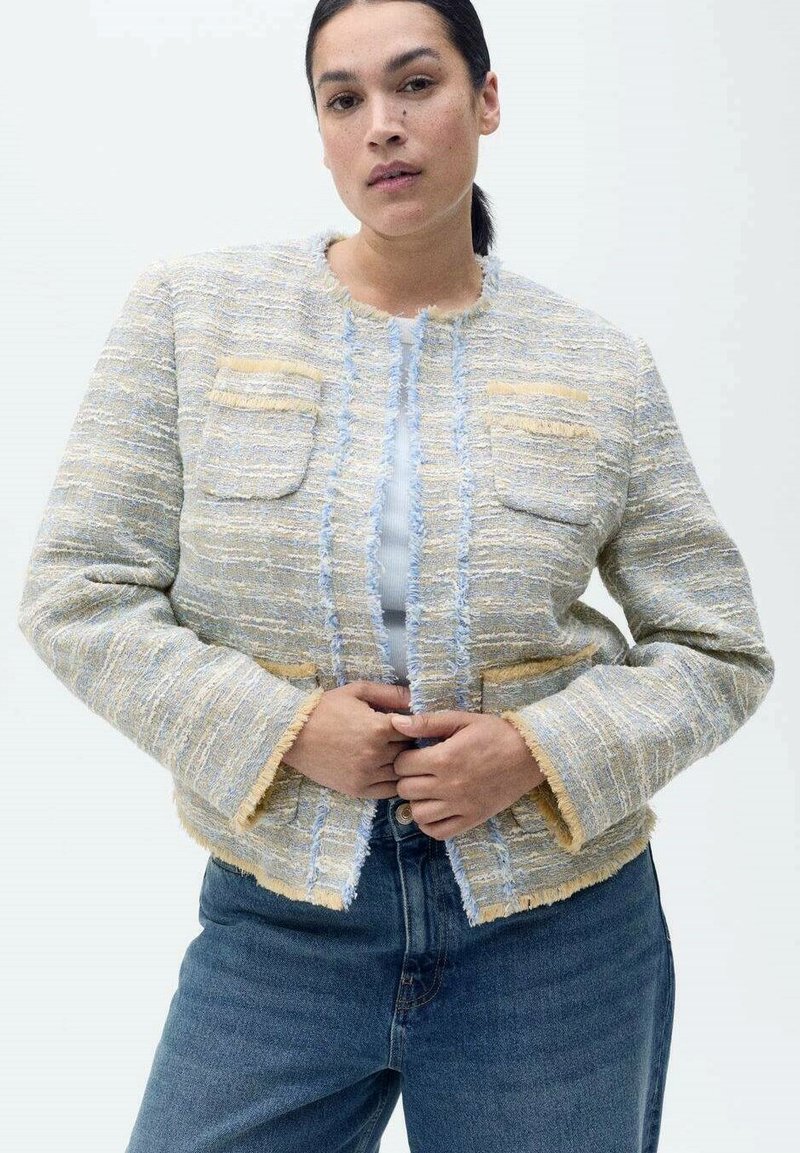 Giacca in tweed blu chiaro e giallo con texture, bordi sfrangiati, due tasche frontali e design cropped, indossata sopra un top blu e dei jeans.