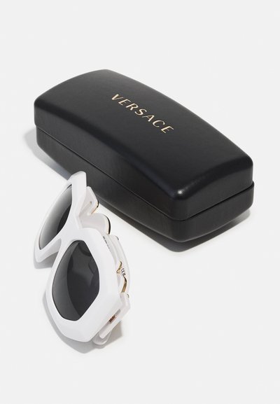 White oversized sunglasses with dark lenses and gold accents, placed next to a black rectangular case featuring "Versace" in gold lettering.