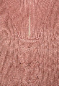 Närbild av en dammig rosa stickad tröja med en vertikal dragkedja och ett strukturerat flätat mönster på framsidan.