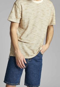 Camiseta de manga corta beige texturizada con cuello redondo, combinada con pantalones cortos de mezclilla oscuros. Decorada con sutiles motas de color marrón claro.