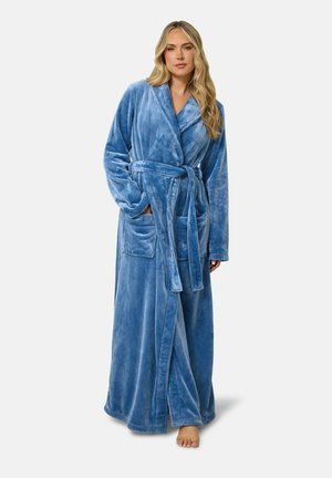 Femme aux longs cheveux blonds portant un peignoir en peluche bleu longue longueur avec poches et ceinture nouée à la taille, debout pieds nus sur fond blanc.