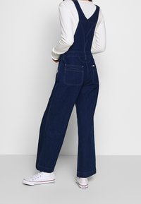 Ciemnoniebieskie dżinsowe ogrodniczki o szerokich nogawkach, z regulowanymi ramiączkami i kieszeniami z tyłu. W komplecie z białą koszulą z długim rękawem i białymi trampkami.