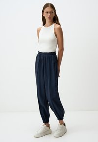 Witte mouwloze top gecombineerd met marineblauwe jogpants met een elastische tailleband en boorden. Model draagt witte sneakers.