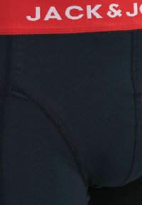 Gros plan d'un sous-vêtement noir avec une large ceinture rouge portant le texte blanc « JACK & J ». Des coutures cousues sont visibles sur le tissu.