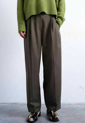 Personne portant un pantalon plissé ample marron foncé, un pull en maille vert et des chaussures à talons à motifs noir, marron et blanc, debout contre un mur uni.