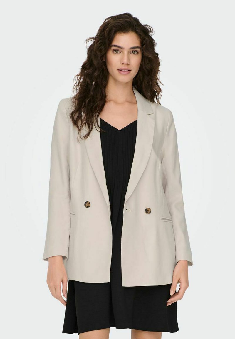 Beige dubbelbreasted blazer van een gladde stof met twee voorzakken en ronde bruine knopen, gecombineerd met een zwarte geplooide jurk.