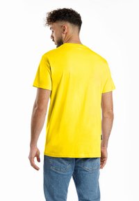 Gelbes Baumwoll-T-Shirt mit kurzen Ärmeln und Rundhalsausschnitt, das eine glatte Textur aufweist und ein Etikett an der unteren Naht trägt, kombiniert mit blauen Jeans.