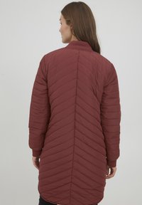 Vadderad kappa i djup röd med zigzag-quilting, långa ärmar och ribbade muddar. Har en hög krage och en längd som går under höfterna.