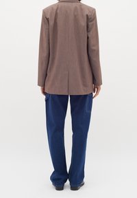 Persona che indossa un blazer marrone e jeans larghi blu, in piedi con la schiena rivolta verso la fotocamera su uno sfondo bianco semplice.