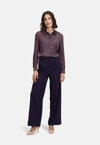 Long-sleeve, patterned shirt in purple and blue, paired with wide-leg navy pants. Model wears light beige heels, hair in loose waves.